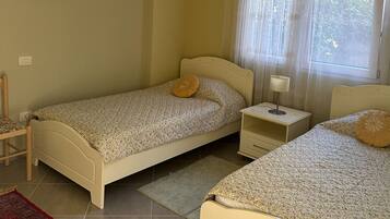 Une chambre avec deux lits simples, une table de chevet avec une lampe, une chaise, un tapis rouge à motifs et une fenêtre avec des rideaux légers.