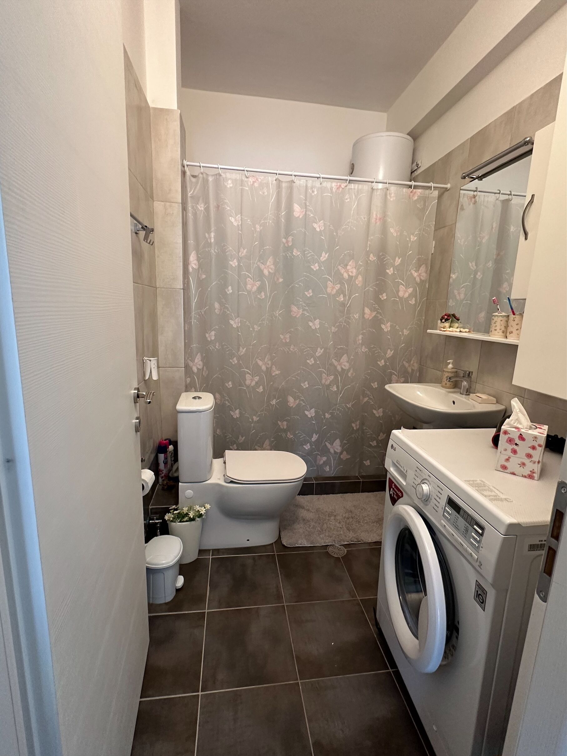 Une petite salle de bain comprenant des toilettes, un bidet et un lave-linge, agrémentée d’un rideau de douche à motifs floraux et d’un lavabo avec un miroir au-dessus.