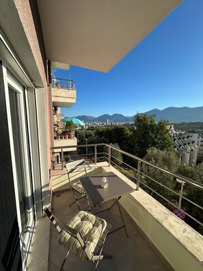 Un balcón con mesa y sillas, con vista a un paisaje urbano y montañas.