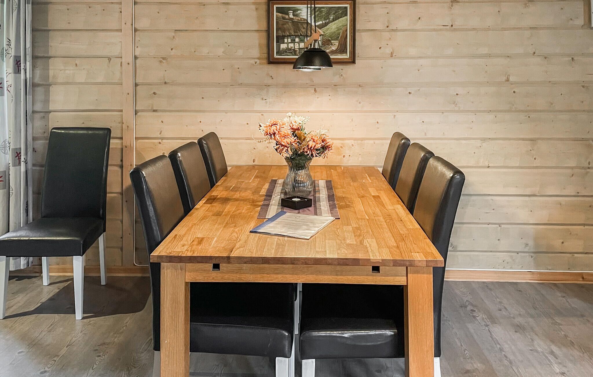 A wooden dining table with six black leather chairs, a vase with flowers, and a framed picture on the wall.