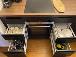 Een keuken met twee open lades vol mokken en servies, een zwart aanrechtblad en een houten snijplank.