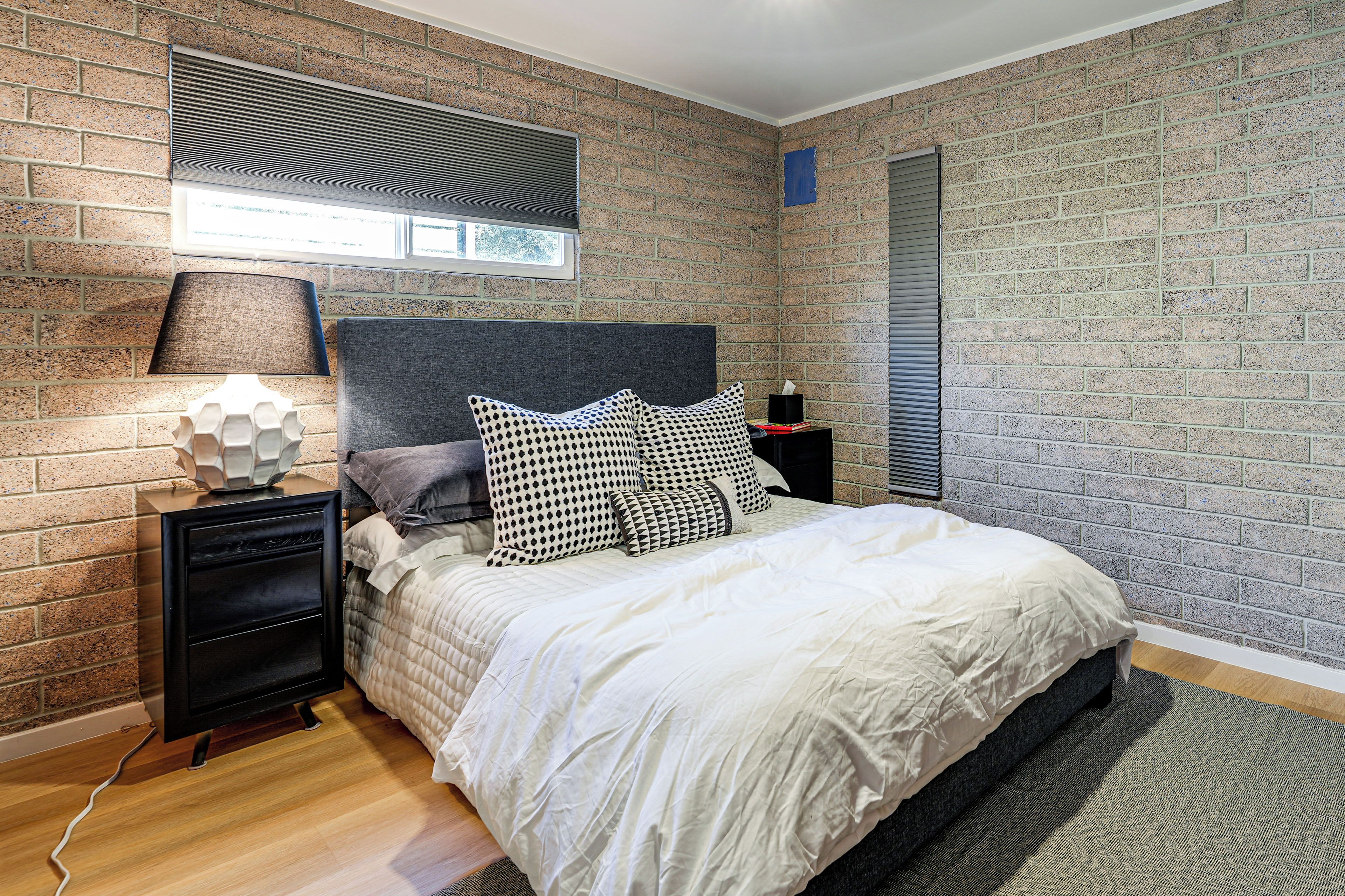 Een slaapkamer met een met baksteen patroon beklede muur, een bed met wit beddengoed, een grijs hoofdeinde en een nachtkastje met een lamp.