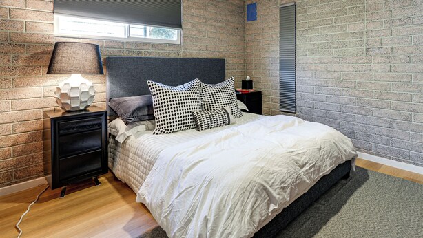 Ein Schlafzimmer mit einer ziegelartigen Wand, einem Bett mit weißer Bettwäsche, einem grauen Kopfteil und einem Nachttisch mit einer Lampe.