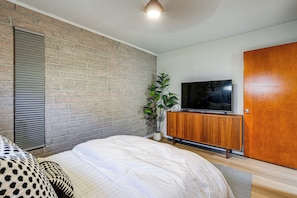 Un dormitorio con pared de ladrillo, cama con edredón blanco, mueble de madera con televisor y puerta de madera.