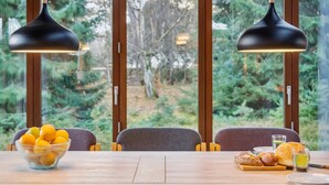 Un comedor con una mesa de madera, dos lámparas colgantes, un cuenco de fruta y vistas a un bosque a través de amplios ventanales.