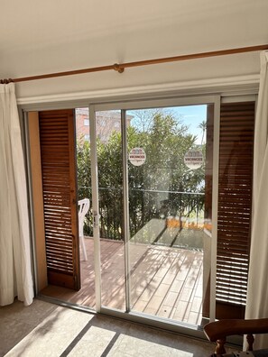 Una stanza con una porta scorrevole in vetro che si apre su uno spazio esterno con un patio in legno e vista su alberi e palazzi.