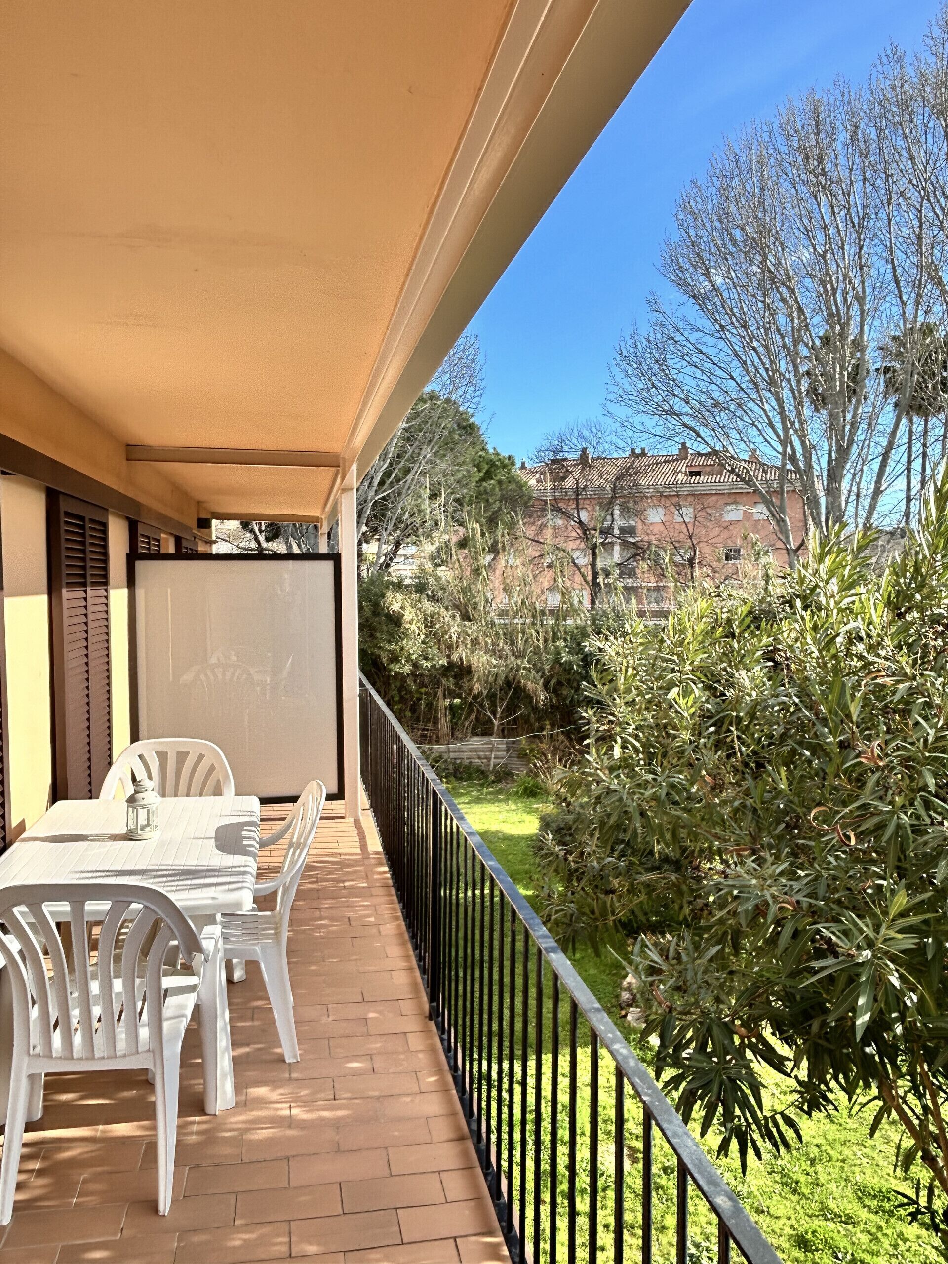 A balcony with a table and chairs, a view of a garden, and a building in the background.