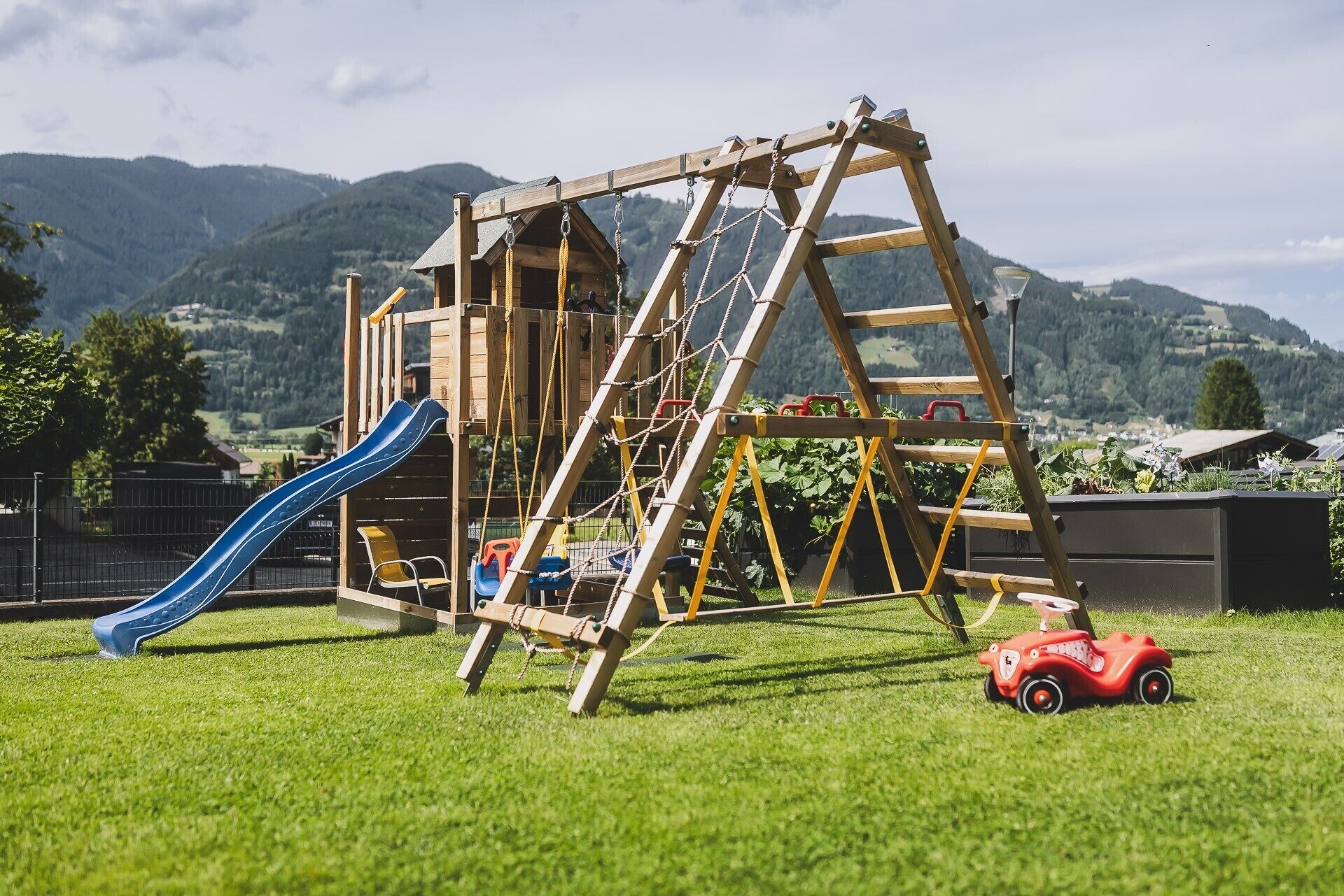 Een houten speelconstructie met een glijbaan en klimnet, voor een bergachtige achtergrond.