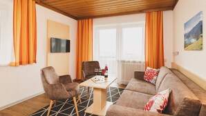 A furnished living room with a wooden ceiling, a television mounted on the wall, a sofa, two chairs, a table with a bottle and glasses, and a window with curtains. - Apartment 16 – Haus Voglreiter with Mountain View, Shared Garden and Wi-Fi (Kaprun)