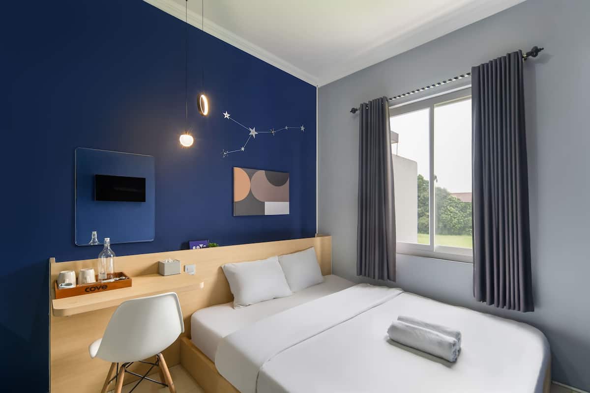 Une chambre d’hôtel comprenant un lit, un bureau avec une chaise, une télévision, une fenêtre avec des rideaux et des œuvres d’art accrochées au mur.