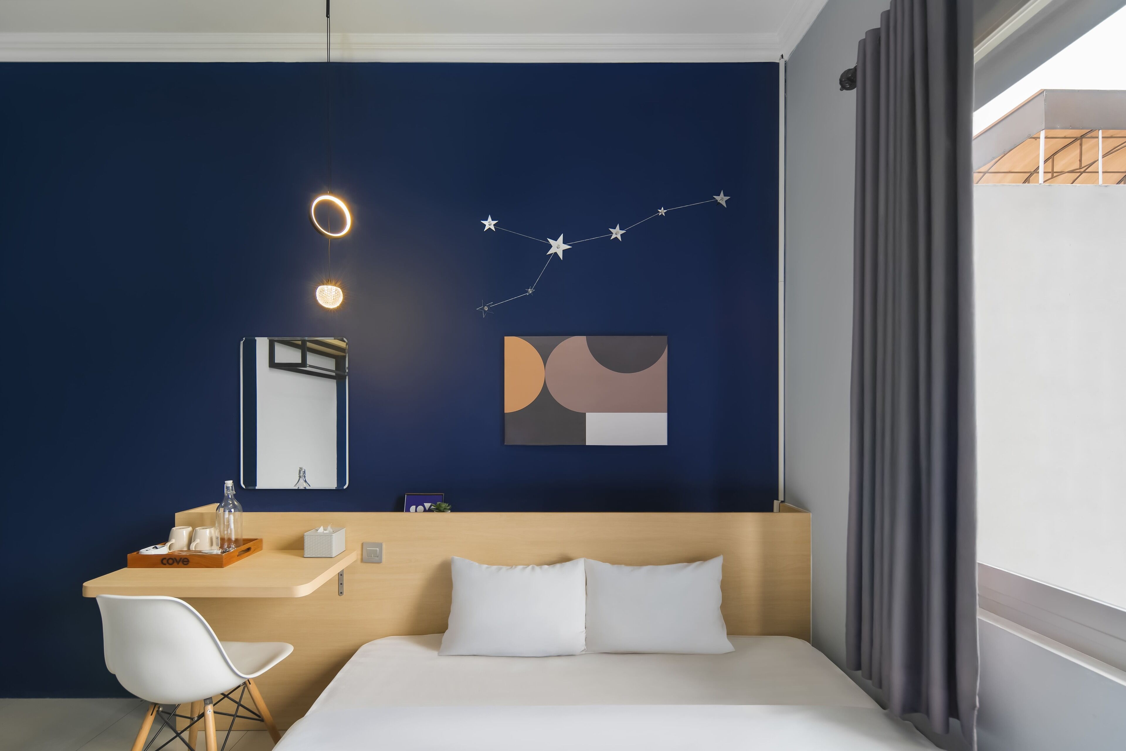 A hotel room with a bed, a desk with a chair, a mirror, and abstract wall art.