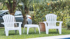 Zwei weiße Stühle und ein kleiner Hocker in Poolnähe, mit einer Palme und einem Auto im Hintergrund.