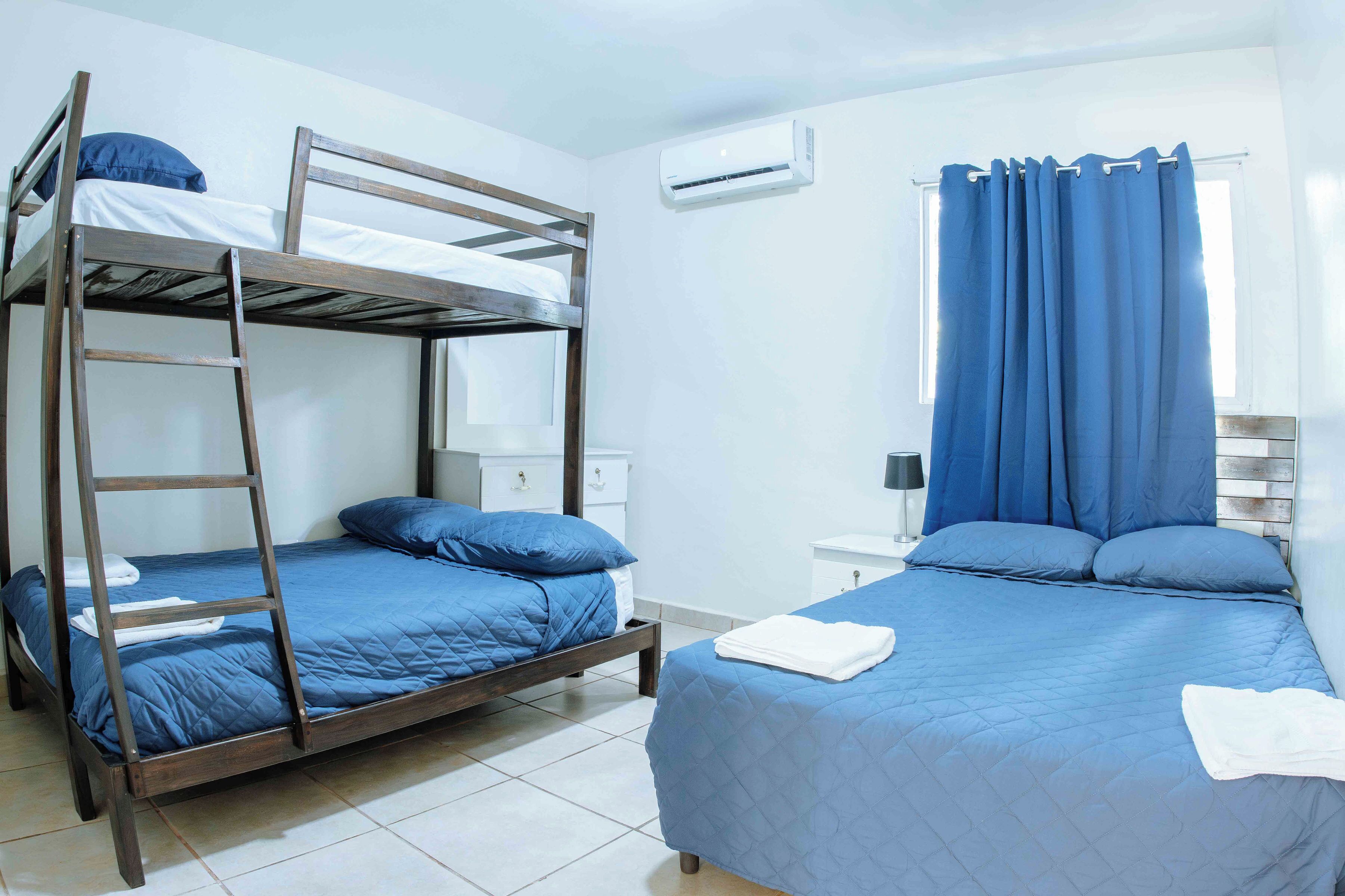 A room with a bunk bed, a single bed, a bedside table, an air conditioning unit, and a window with curtains.