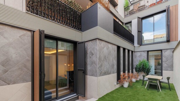 Un edificio moderno con puerta de entrada de vidrio, un pequeño espacio exterior con mesa y sillas, y un césped bien cuidado.
