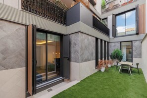 A modern building with a glass door entrance, a small outdoor seating area with a table and chairs, and a well-maintained lawn. - w* | Comfy 2BR w/ Great Terrace in Chueca (Madrid)