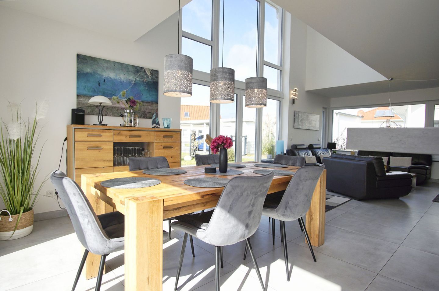 A modern dining area with a wooden table and grey chairs, a kitchen with a large window, and a living area with a sofa and a coffee table.