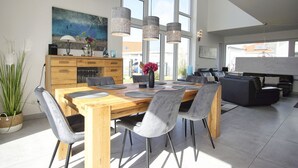 A modern dining area with a wooden table and grey chairs, a kitchen with a large window, and a living area with a sofa and a coffee table. - Villa Kristall - house in an idyllic location with conservatory, terrace and garden (Trent)
