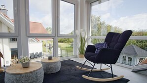 A modern living room with large windows, a black rocking chair, two round side tables, and a potted plant. - Villa Kristall - house in an idyllic location with conservatory, terrace and garden (Trent)
