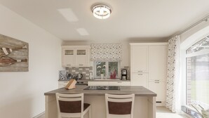 A modern kitchen with a central island, two bar stools, white cabinetry, a coffee machine, and a window with curtains.