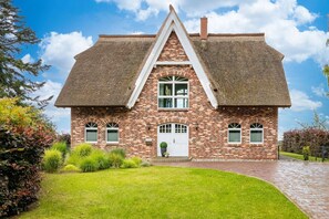 A brick house with a steeply pitched thatched roof, a central entrance with a white door, and a small front garden.