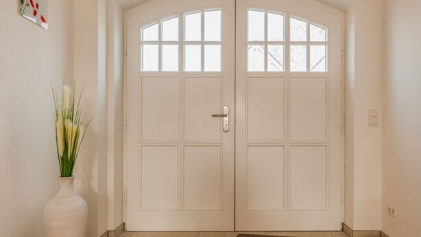 Eine weiße Doppeltür mit Glasfenster, eine Fußmatte und ein Blumenvasen mit Grünpflanzen auf dem Boden.