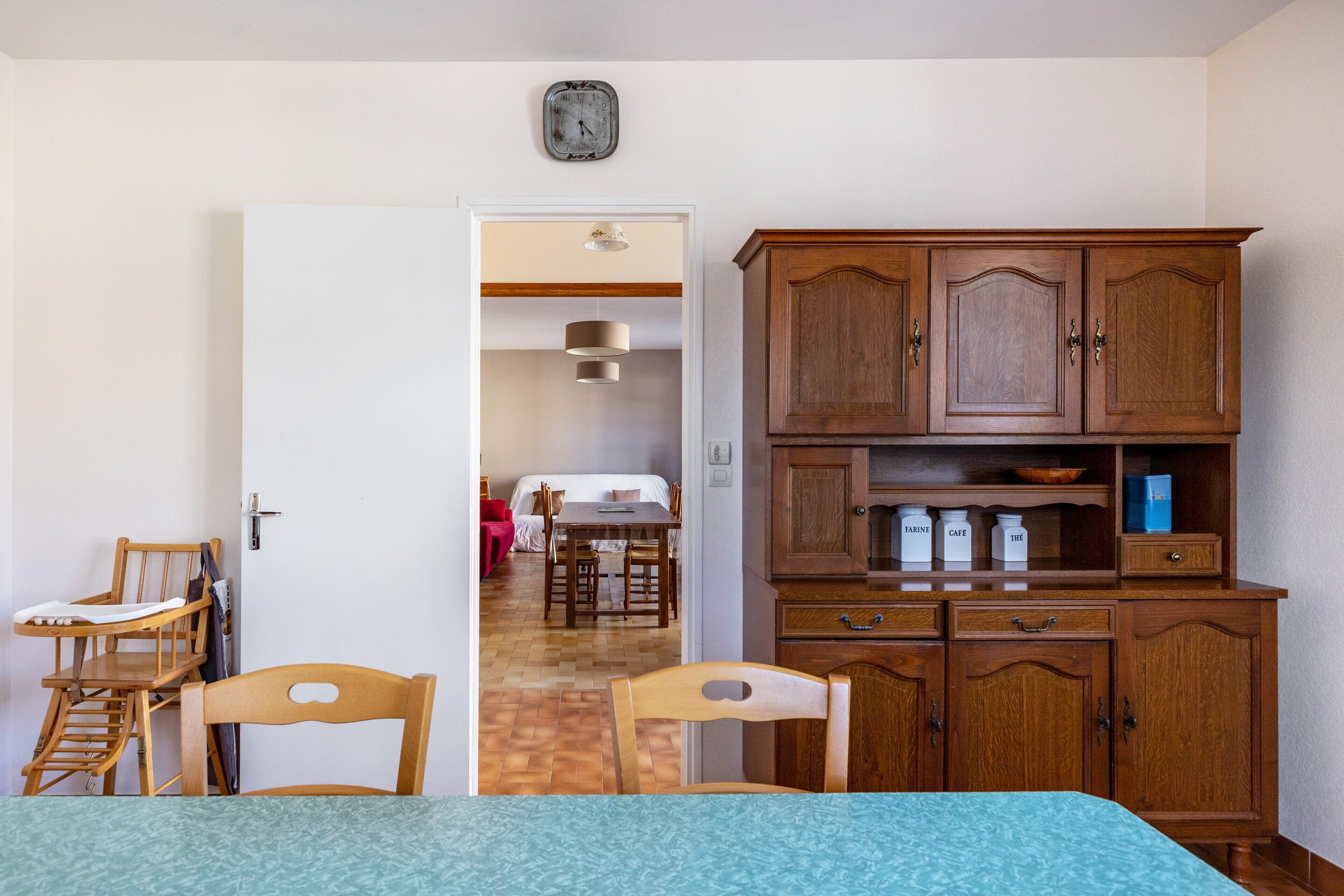 A room with a bed, wooden chairs, a dining table, a wooden cabinet, and a clock on the wall.