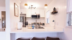 A modern kitchen with a central island, bar stools, a microwave, and a coffee machine. - Ocean+Mountain+Pool View Upgraded Condo- Steps from the beach centrally located (Kapaʻa)