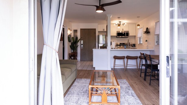 A modern kitchen with white cabinets, a microwave, and a coffee machine. There is a dining area with a glass table and wooden chairs. - Ocean+Mountain+Pool View Upgraded Condo- Steps from the beach centrally located (Kapaʻa)