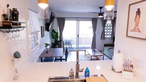 A modern kitchen with a sink, faucet, and countertop. There are wine glasses on a shelf, a plant, and a dining area with a table and chairs. - Ocean+Mountain+Pool View Upgraded Condo- Steps from the beach centrally located (Kapaʻa)