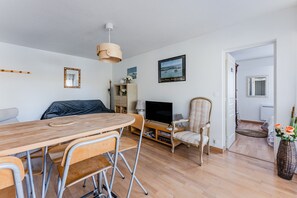 Un salon comprenant une table à manger en bois, des chaises, un téléviseur, une bibliothèque et un fauteuil avec un coussin.