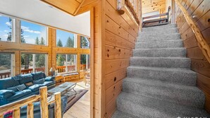 Interior de una cabaña de madera con una escalera que conduce a un balcón, ventanas amplias y una zona de estar acogedora.