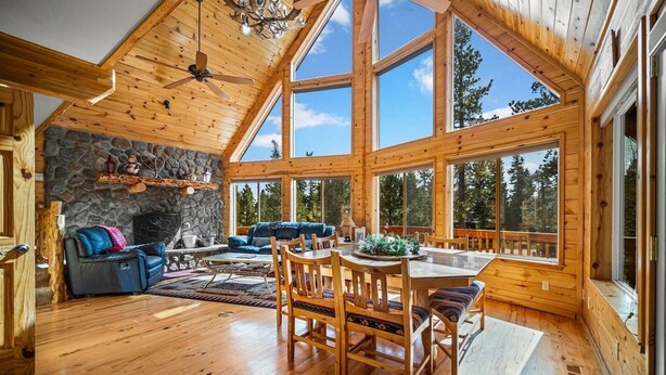 Una cabaña de madera espaciosa con comedor, chimenea de piedra y ventanas amplias que dan vistas al bosque.