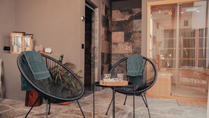 Ein Zimmer mit zwei schwarzen Stuhlen mit Drahtgestell, einem kleinen Holztisch mit Gläsern und einer Serviertablett sowie einem steinbelagten Boden.