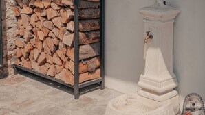 Ein rustikaler Innenraum mit Steinboden, einem Holzstoß, einem weißen Podest mit Wasserhahn und einer kleinen Igel-Figur.