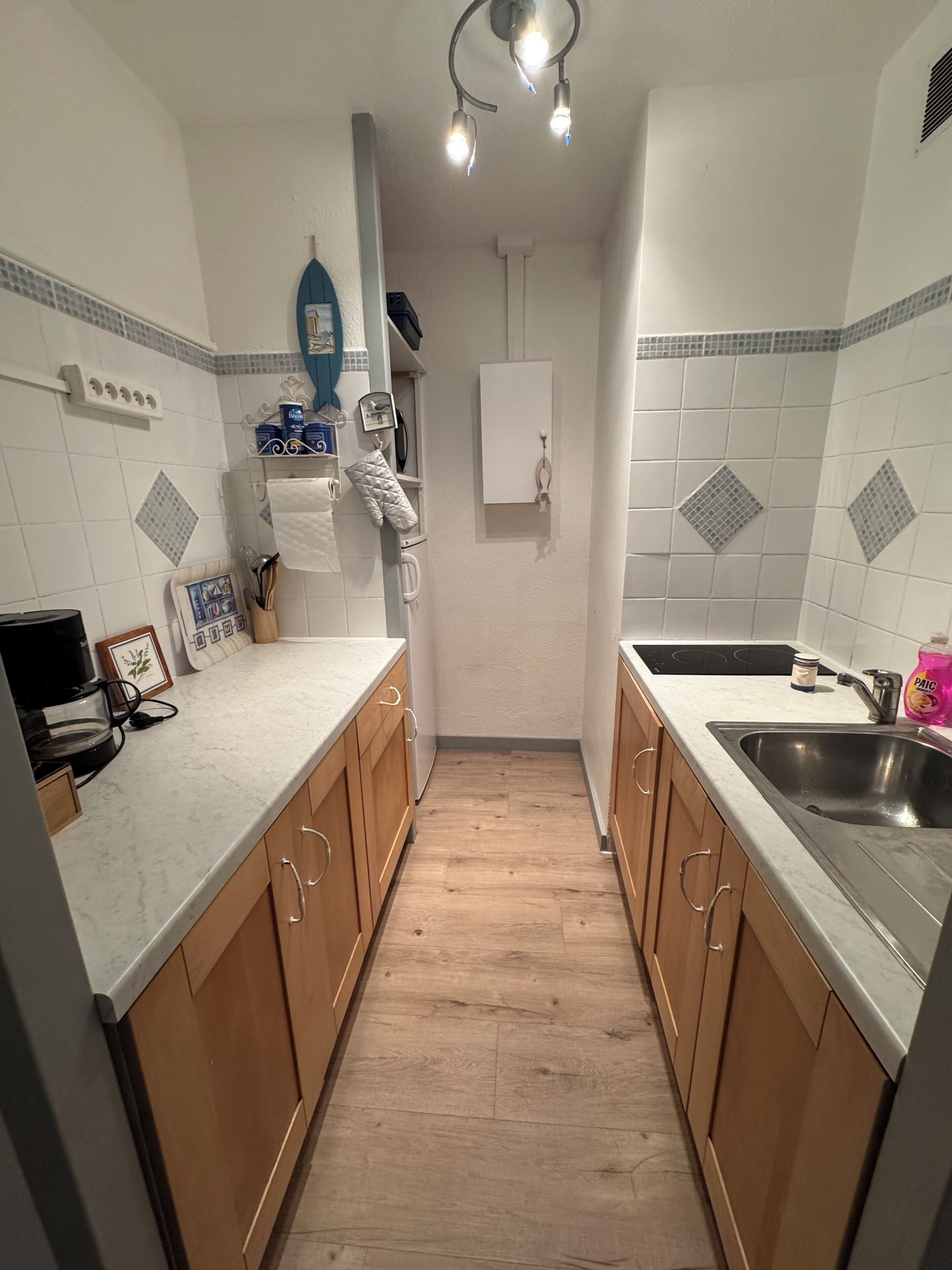 Eine schmale Küche mit Holzschränken, einer Edelstahl-Spüle und einer weißen Arbeitsplatte.