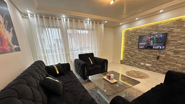 A modern living room with black sofas, a glass coffee table, and a stone accent wall with a TV. - Stylish apartment in a modern ambience in Tuttlingen (Tuttlingen)