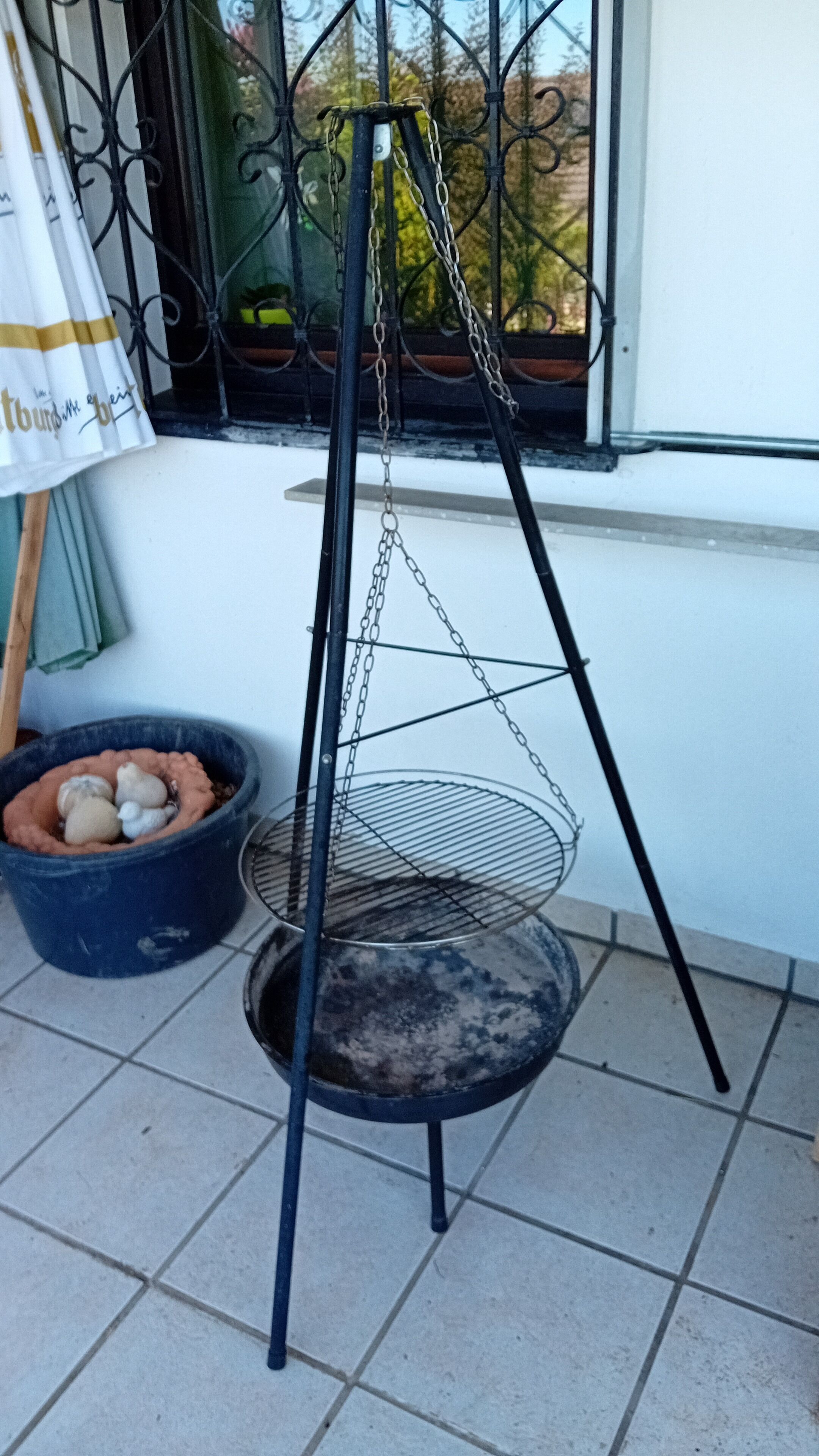 A metal hanging basket with a chain, placed on a tiled floor next to a window with a metal grill.