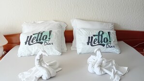 Una cama bien hecha con dos almohadas que dicen “¡Hola, querida!” y dos toallas dobladas sobre la cama.