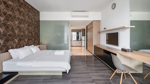 A modern hotel room with a large bed, a desk with a chair, a TV, and a kitchen area in the background. - Cove Southouse Residence (Jakarta)