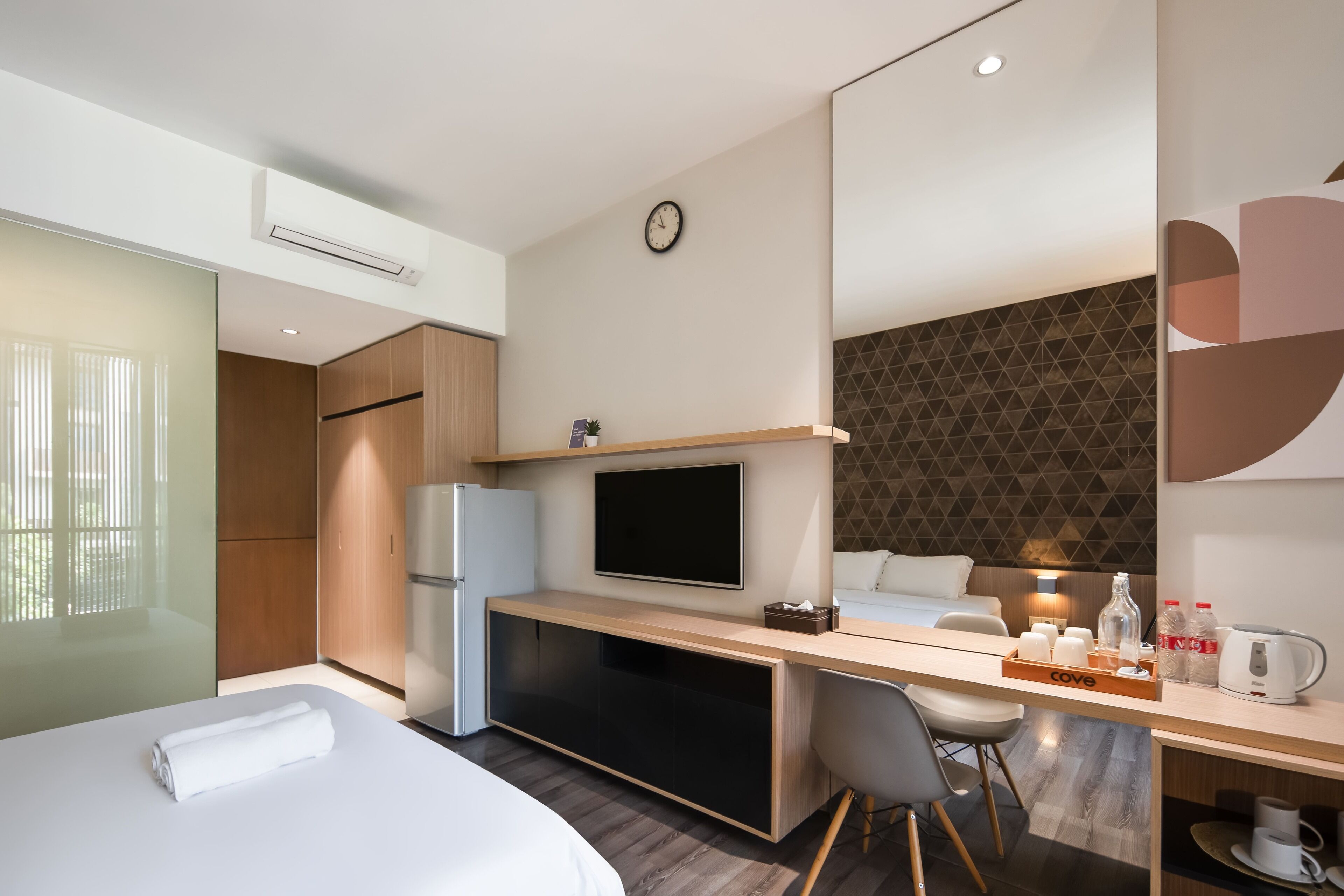 A modern hotel room with a bed, a desk with a computer, a television, and a small kitchenette.