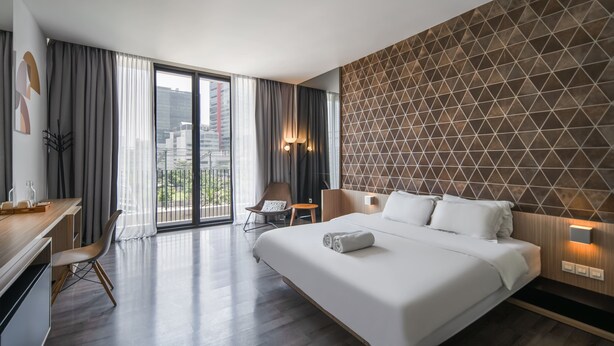 A modern hotel room with a large bed, a desk, a chair, and a view of the city through large windows. - Cove Southouse Residence (Jakarta)