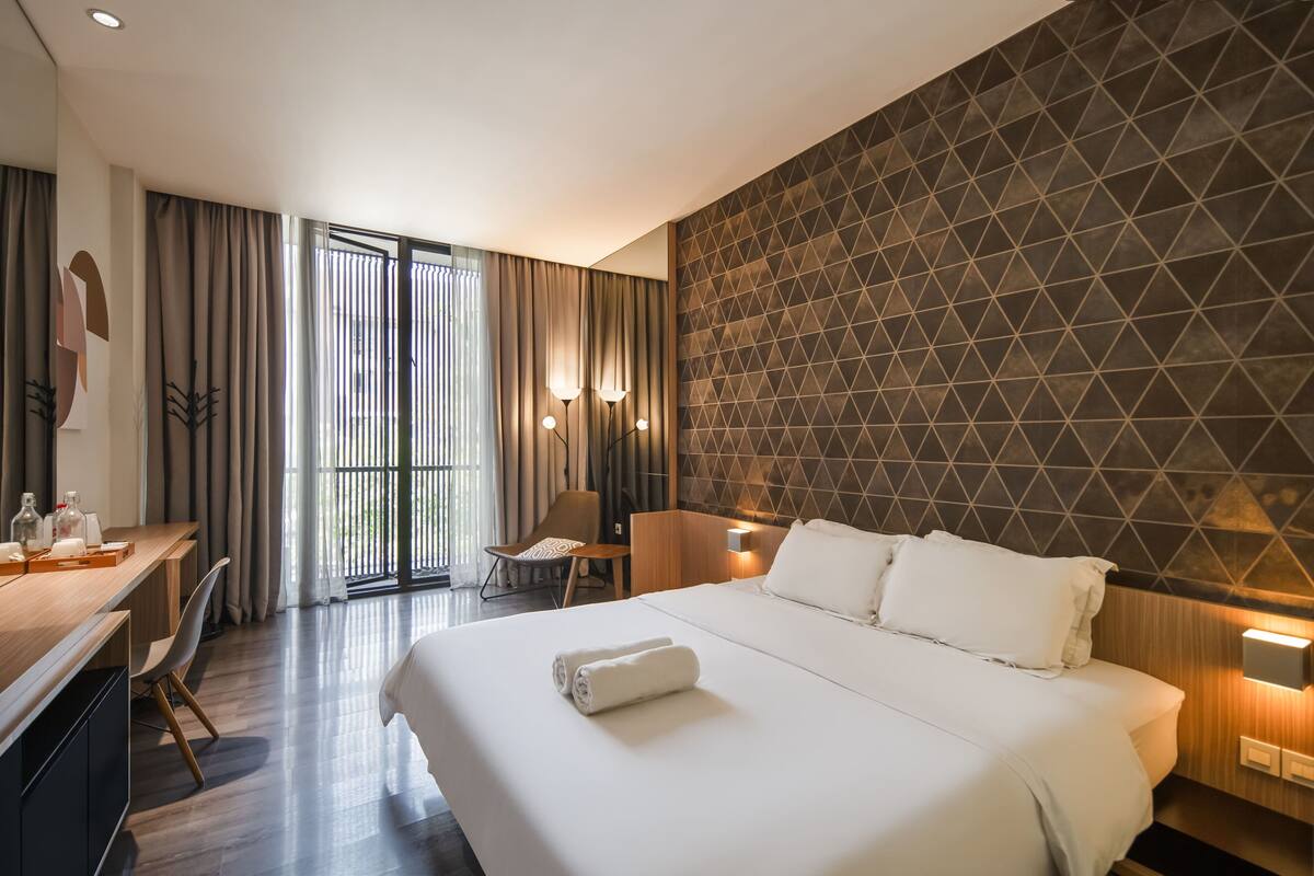 A modern hotel room with a large bed, a desk, and a chair. There is a view of the outdoors through the window.