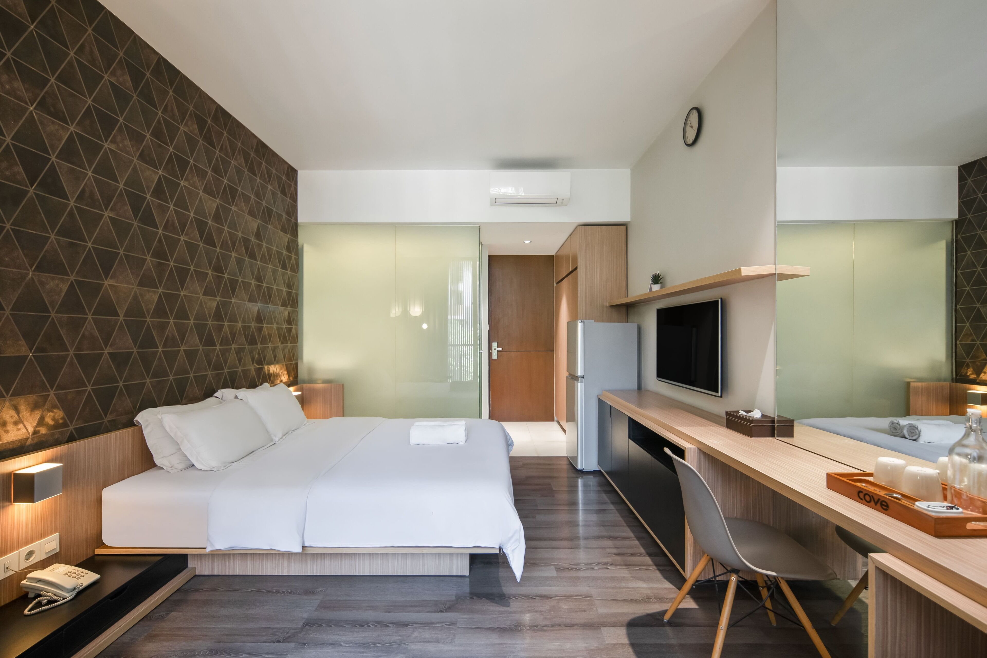 A modern hotel room with a large bed, bedside table, TV, and a desk with a chair.