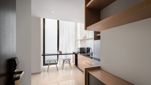 A modern kitchen with a black countertop, wooden cabinets, and a stainless steel refrigerator. There is a window with sheer curtains and a view of the outside. - Cove Southouse Residence (Jakarta)