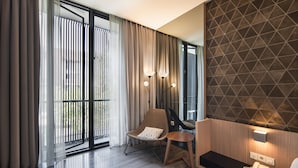 A modern hotel room with a large sliding glass door, a comfortable chair, a small wooden table, and a wall-mounted telephone. - Cove Southouse Residence (Jakarta)