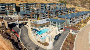 A modern multi-story hotel with a swimming pool, parking area, and a hilly backdrop. - All-Season Escape | Ski-In/Out + Hot Tub + Clubhouse Amenities Canyons Village (Park City)