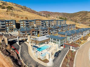 A modern multi-story hotel with a swimming pool, parking area, and a hilly backdrop.