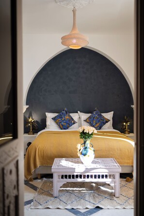 Une chambre à coucher avec un grand lit, une table de chevet blanche, une petite table sur laquelle est posé un vase de fleurs et un luminaire suspendu.