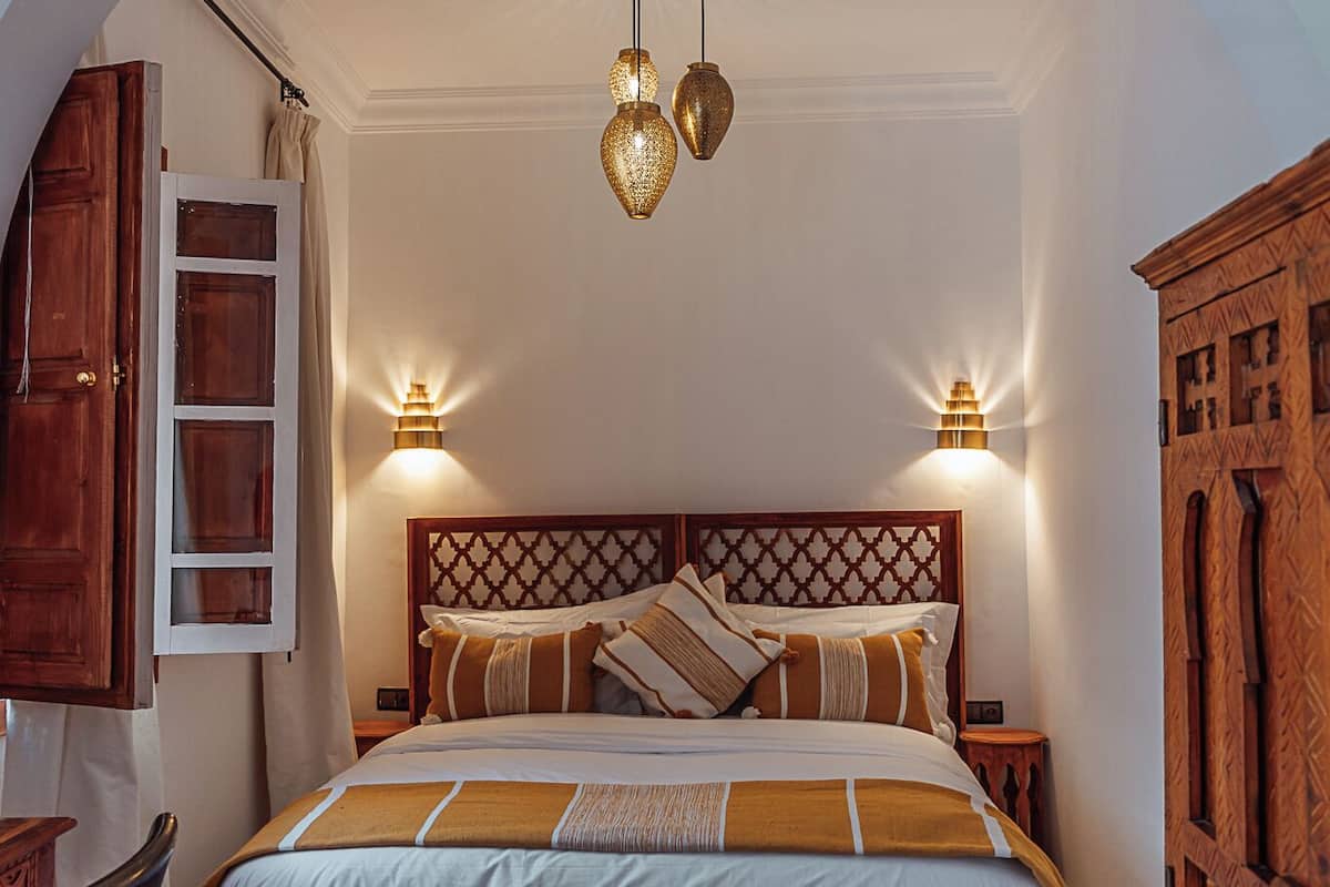 A bedroom with a large bed, wooden headboard, two wall-mounted lamps, a bench, and a wooden wardrobe.