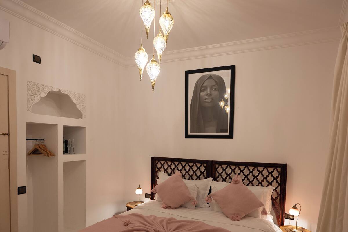 A bedroom with a large bed, a headboard with a geometric pattern, a ceiling light fixture, a framed portrait on the wall, and a bedside table with a lamp.